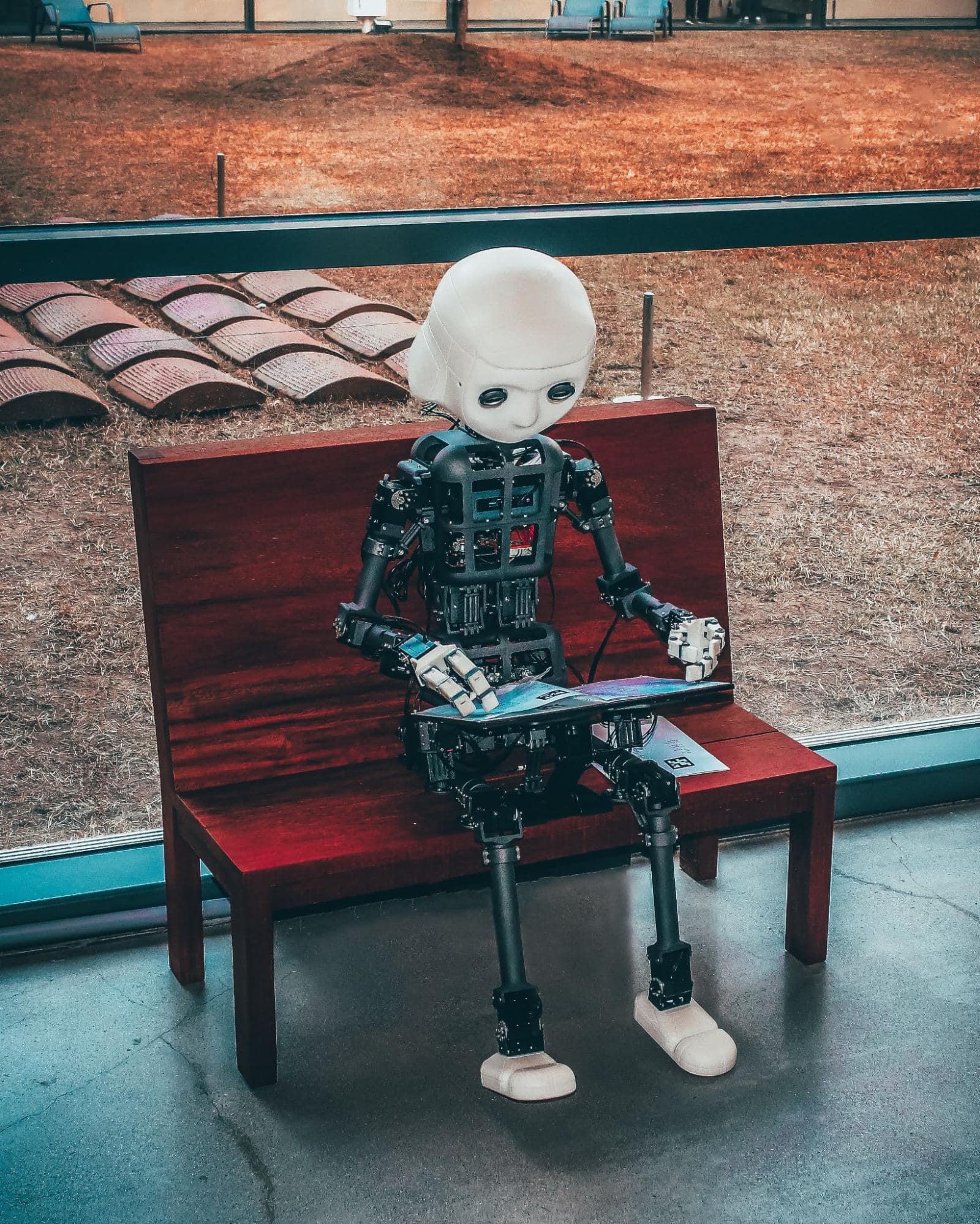 Solemn-looking robot sitting on a bench while holding a laptop